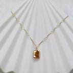 Collier "Ma voie" avec pendentif rectangulaire en acier doré, vue de dos personnalisable sur fond beige texturé.