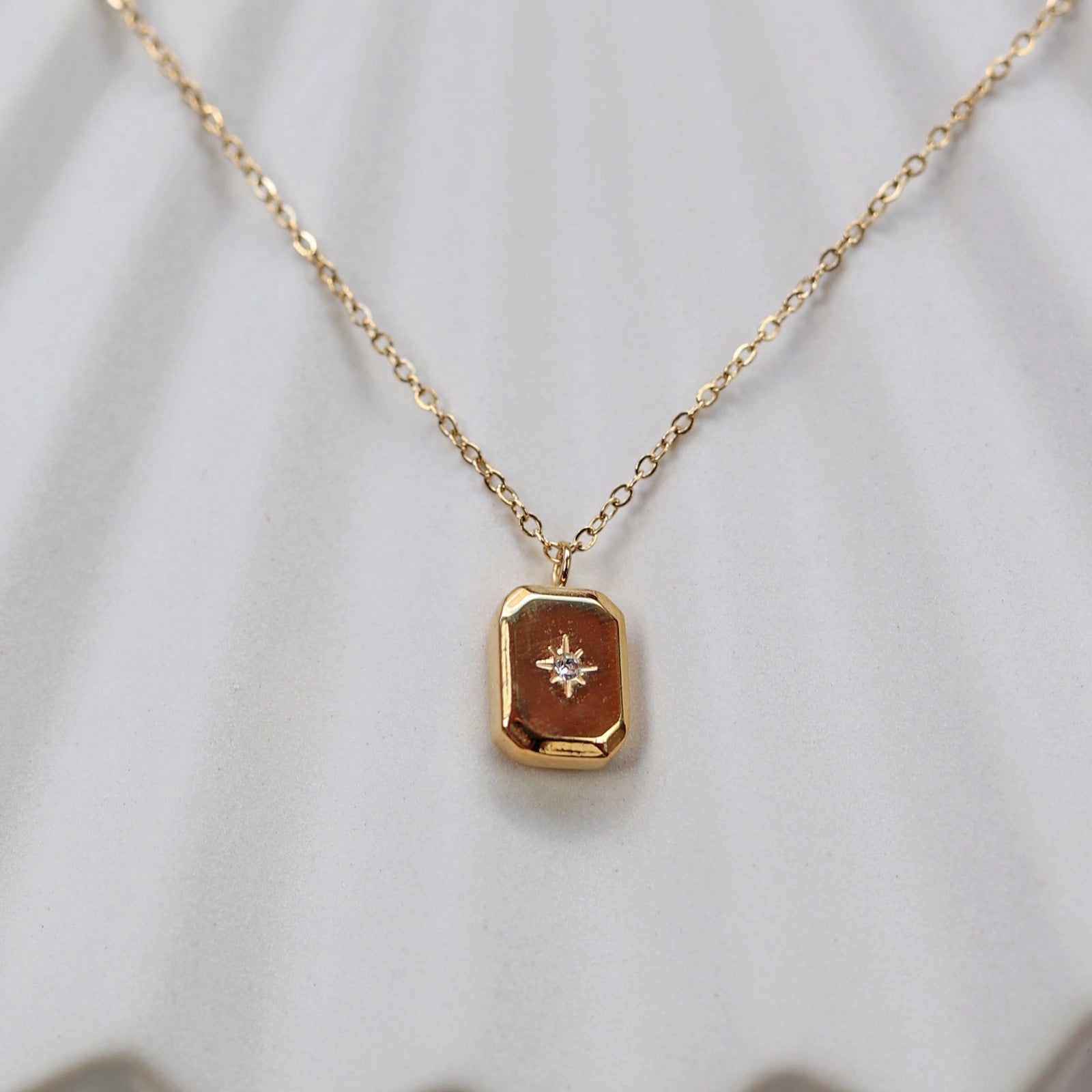 Collier "Ma voie" avec pendentif rectangulaire personnalisable en acier doré, sur fond beige texturé.