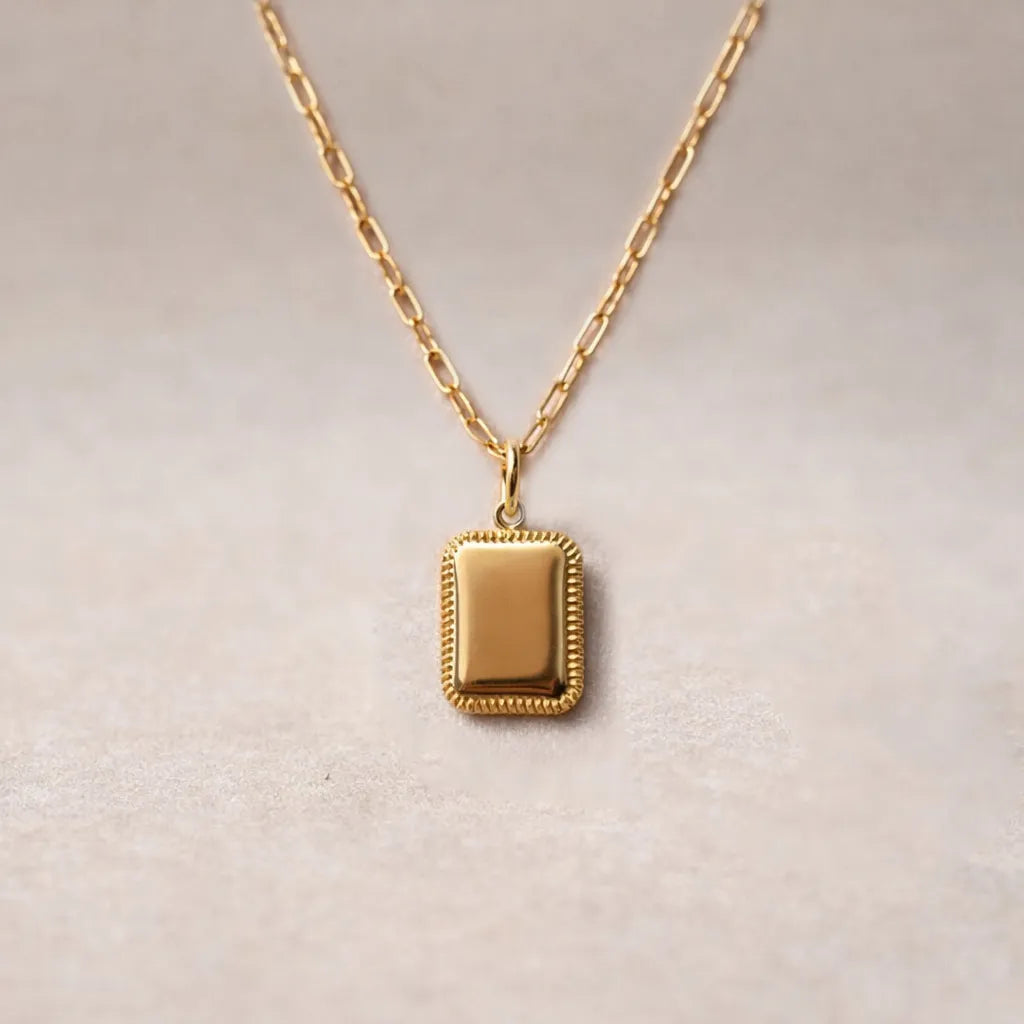 Collier "Ma vérité" avec pendentif rectangulaire personnalisable en acier doré, présenté sur un fond beige texturé.