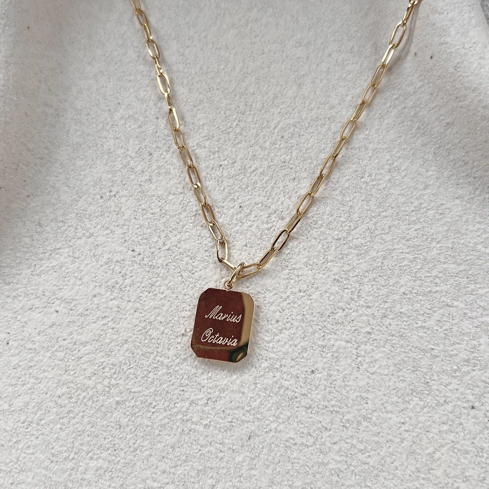 Collier Trésor avec pendentif rectangulaire en acier doré, personnalisé au verso de 2 prénoms présenté sur un fond beige texturé.