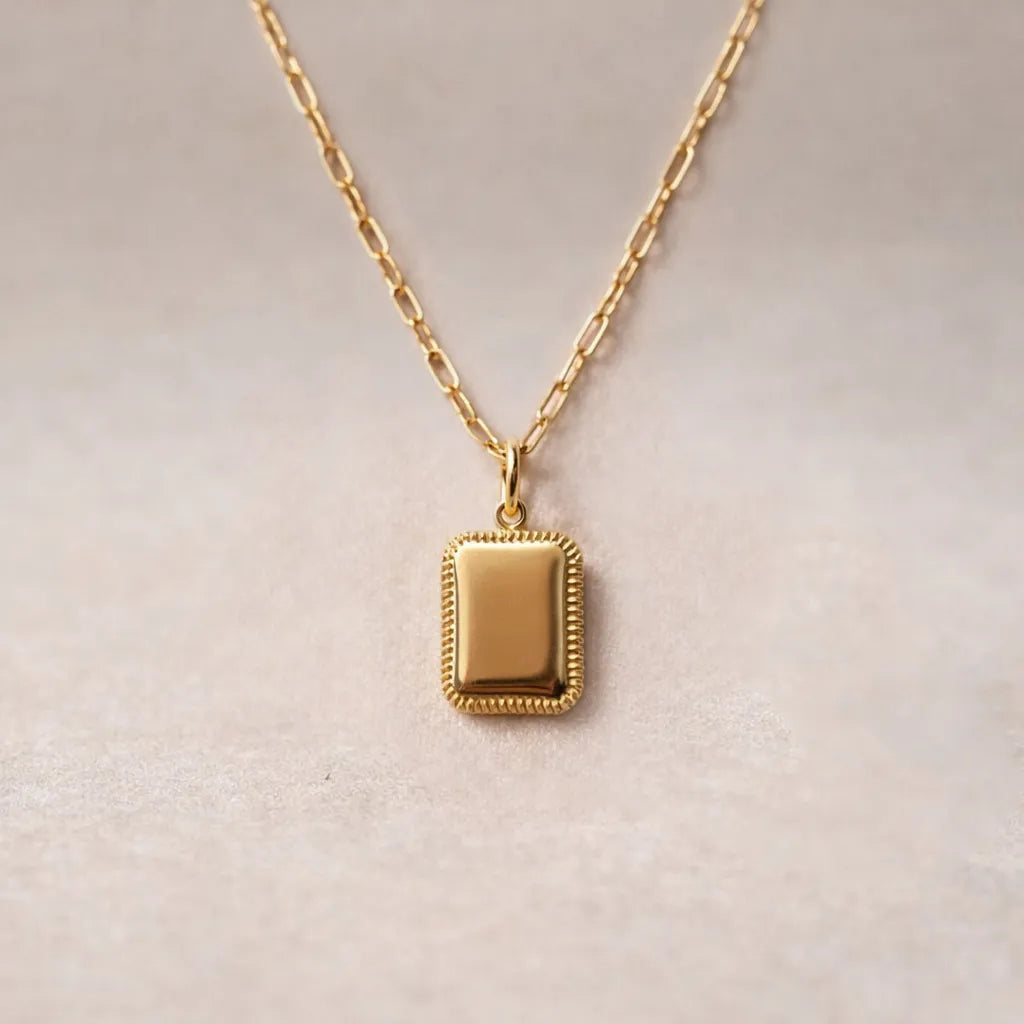 Collier Trésor avec pendentif rectangulaire personnalisable en acier doré, présenté sur un fond beige texturé.