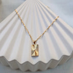 Collier Résilience en acier doré avec pendentif rectangulaire gravé de rayons, personnalisable au verso, vue de face sur support beige texturé - Visée Bijoux