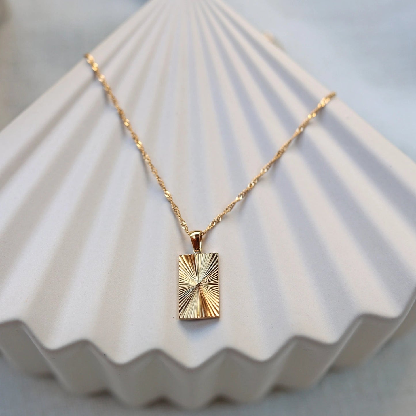 Collier Résilience en acier doré avec pendentif rectangulaire gravé de rayons, personnalisable au verso, vue de face sur support beige texturé - Visée Bijoux
