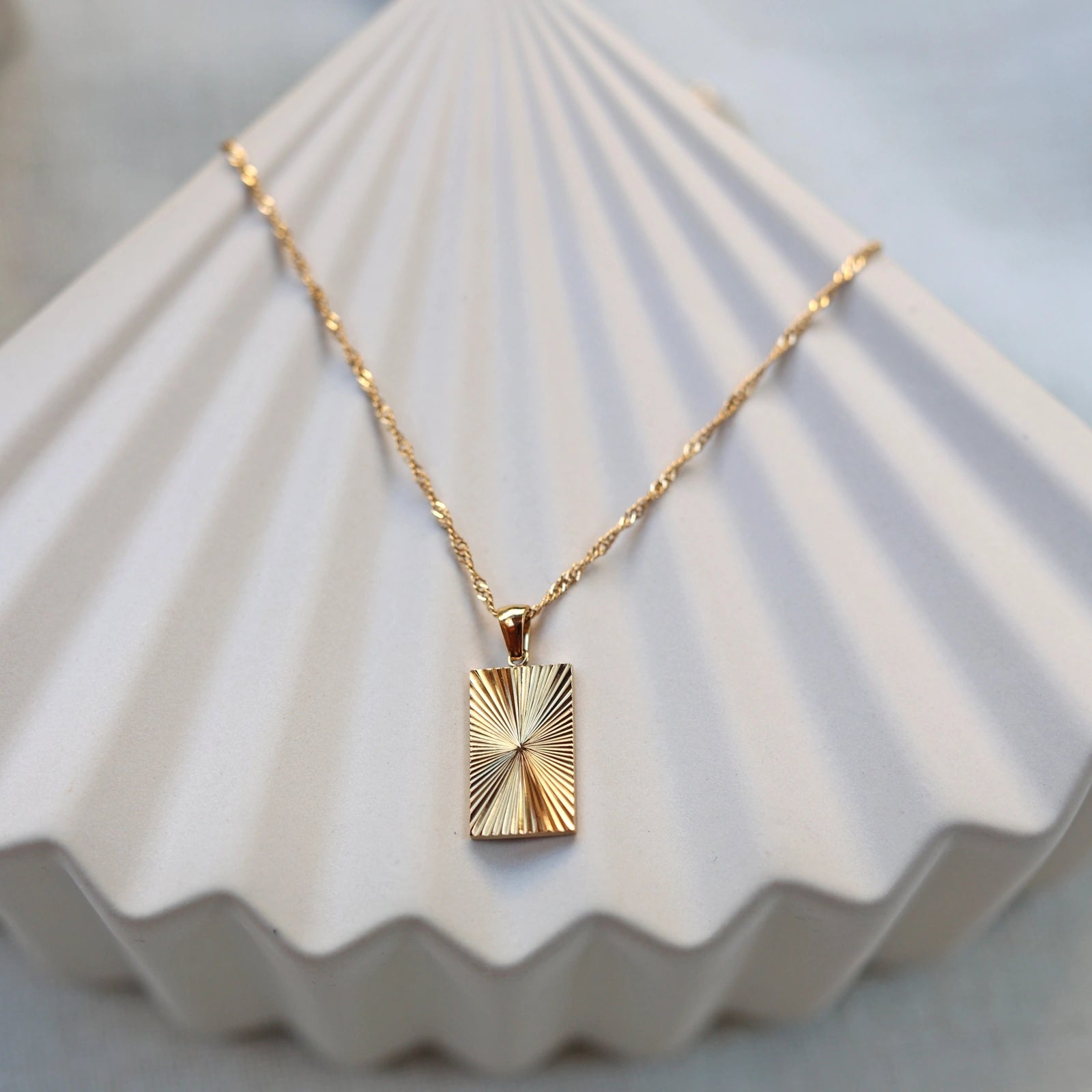 Collier Résilience en acier doré avec pendentif rectangulaire gravé de rayons, personnalisable au verso, vue de face sur support beige texturé - Visée Bijoux