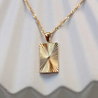 Zoom sur le collier Résilience en acier doré avec pendentif rectangulaire gravé de rayons, personnalisable au verso, vue de face sur support beige texturé.
