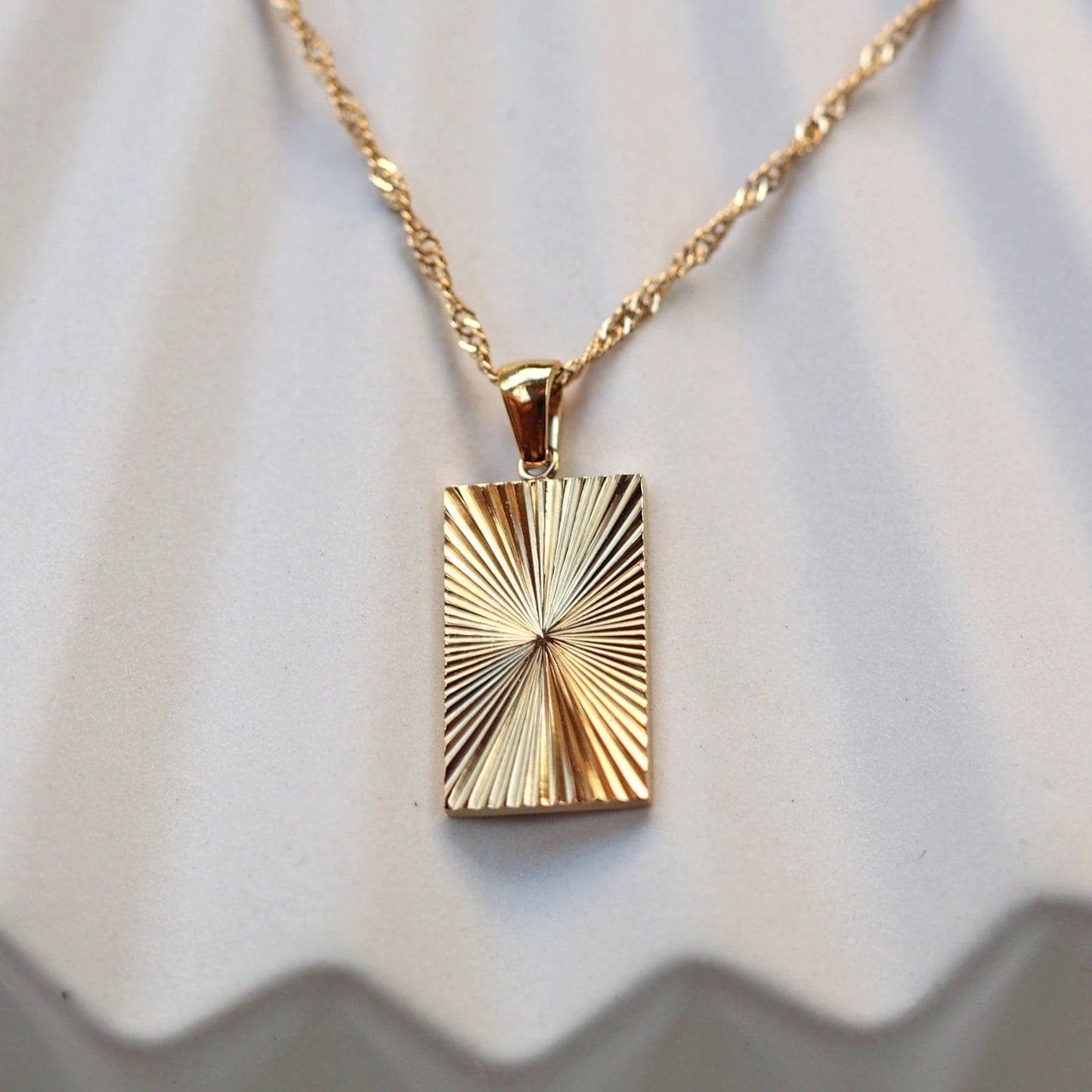 Zoom sur le collier Résilience en acier doré avec pendentif rectangulaire gravé de rayons, personnalisable au verso, vue de face sur support beige texturé.