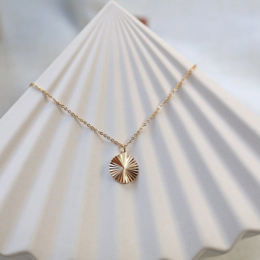 Vue de face du collier "Energie", pendentif rond strié personnalisable en acier doré, posé sur une structure beige.