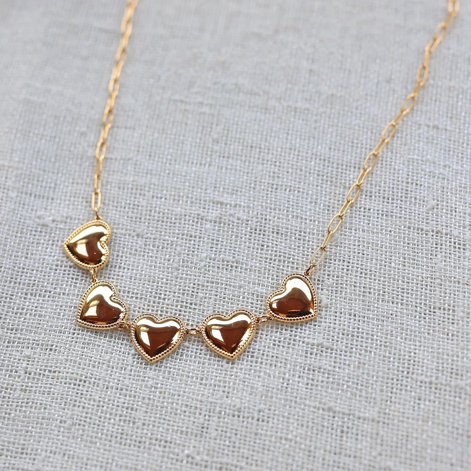 Zoom sur le collier "Je suis là" de Visée, composé de cœurs personnalisables en acier doré, posé sur un fond beige texturé.
