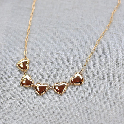 Zoom sur le collier "Je suis là" de Visée, composé de cœurs personnalisables en acier doré, posé sur un fond beige texturé.