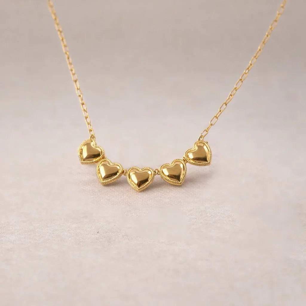 Zoom sur le collier "Je suis là" de Visée, composé de cœurs personnalisables en acier doré, posé sur un fond beige texturé.