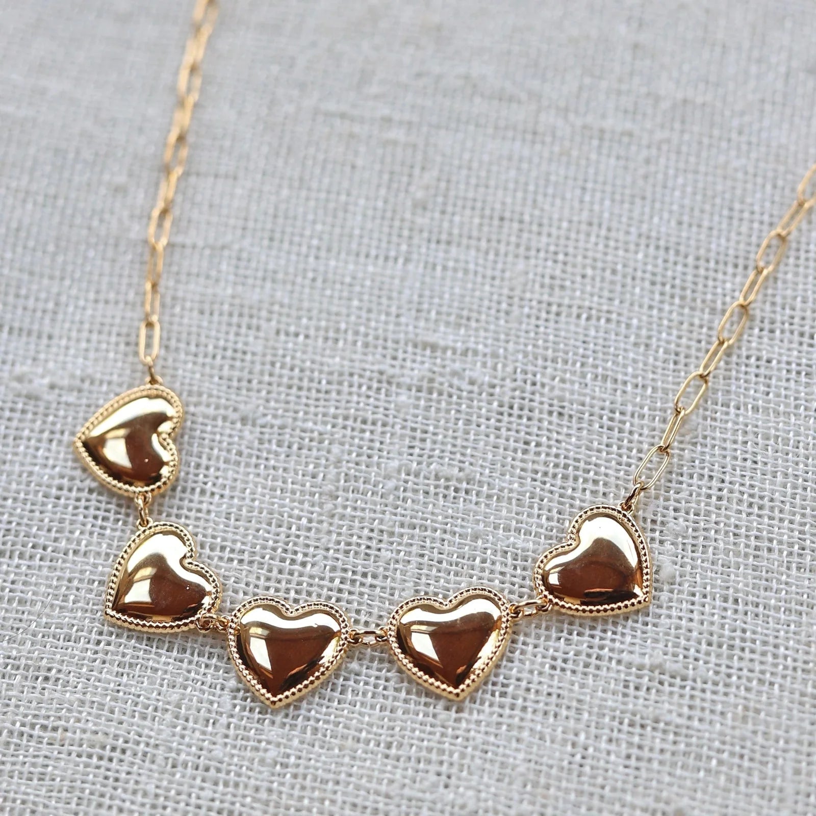 Zoom sur le collier "Je suis là" de Visée, composé de cœurs personnalisables en acier doré, posé sur un fond beige texturé.