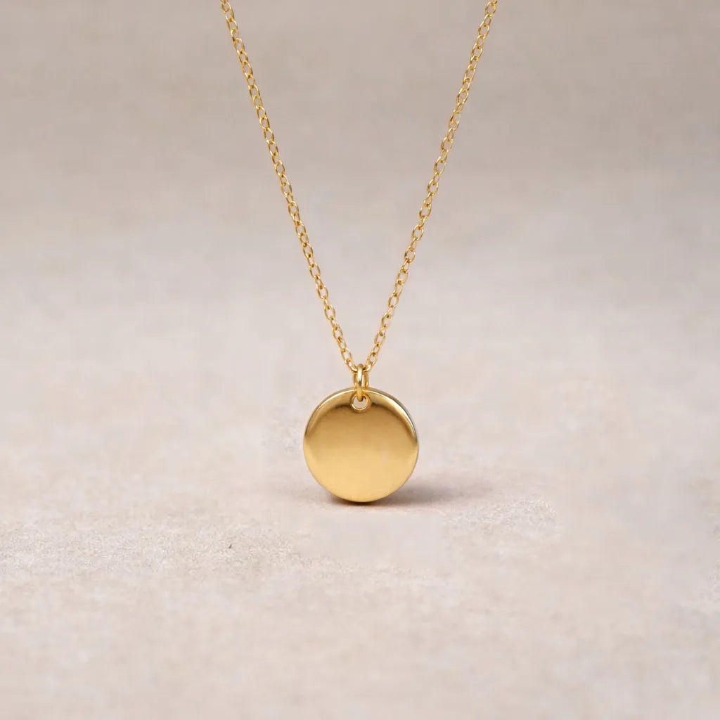 Zoom sur le collier personnalisable "Lumineuse" avec médaille ronde brillante en acier dorée vue de dos, posé sur un fond beige texturé.