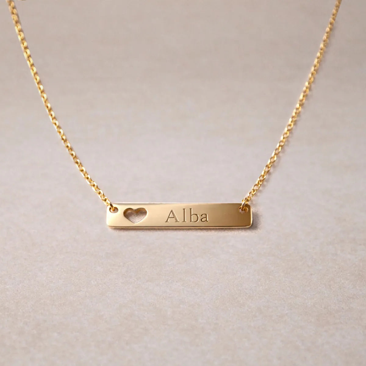 Collier doré personnalisable cœur gravé au nom de Alba – Douceur Visée Bijoux, posé sur fond beige texturé
