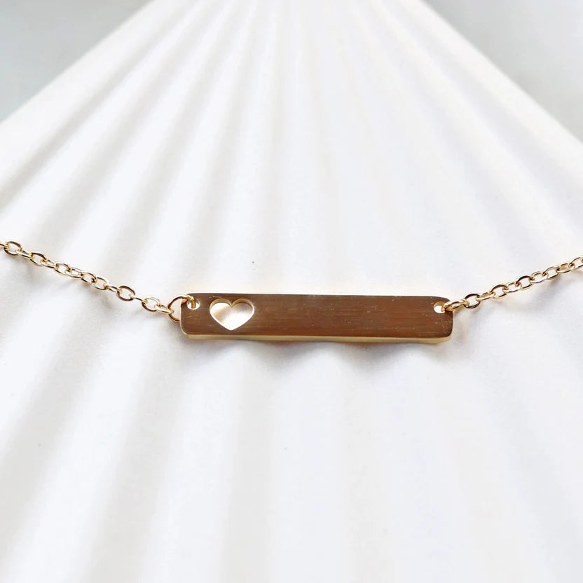 Zoom sur le collier "Touchante" avec plaque personnalisable en acier doré avec cœur découpé, posé sur un support beige texturé.