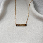 Collier "Touchante" avec plaque personnalisé d'un prénom "Alba" en acier doré avec cœur découpé, posé sur un support beige texturé.