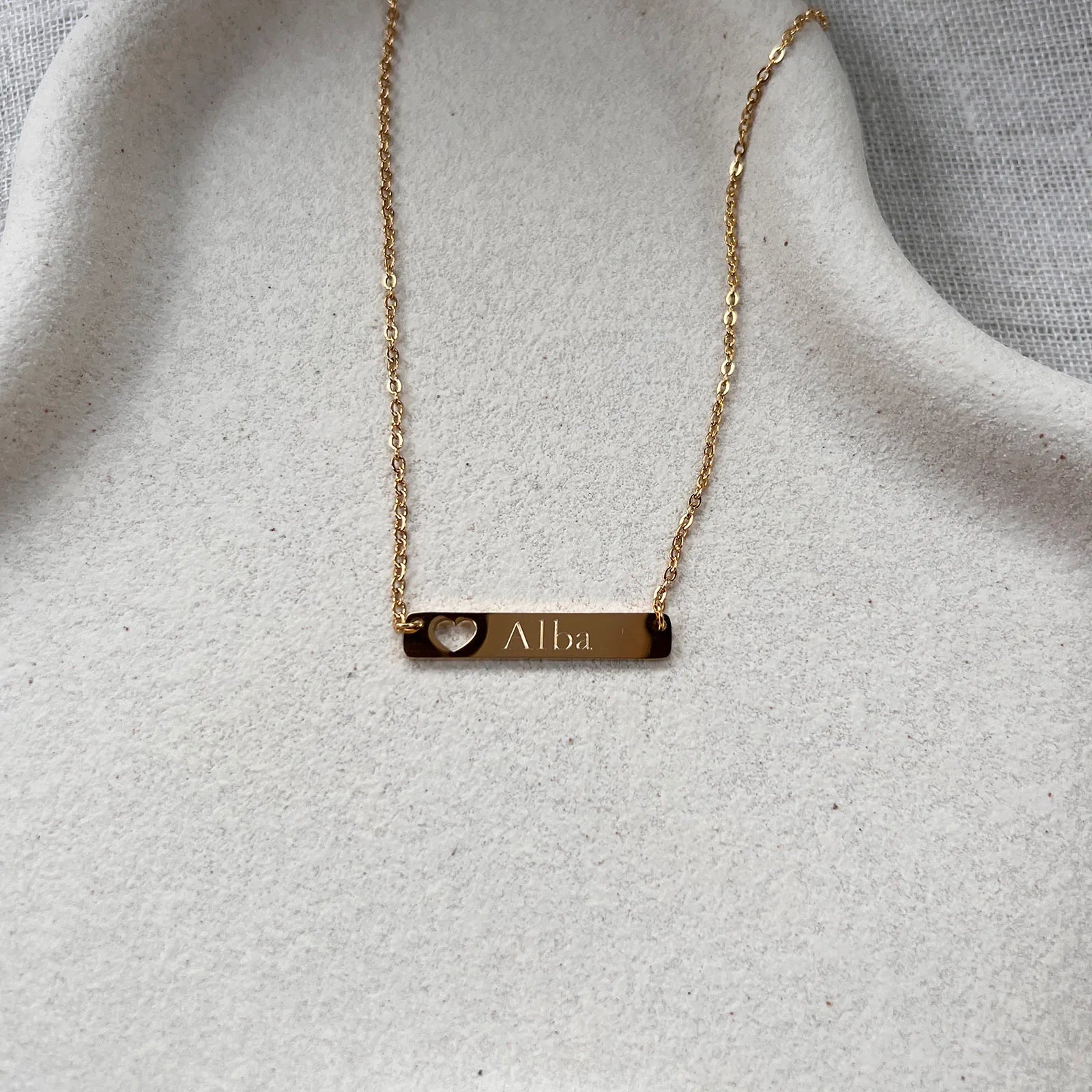 Collier plaque en acier doré gravé d'un prénom, avec cœur découpé , présenté à plat sur un supporte texturé beige. 