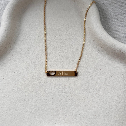 Collier plaque en acier doré gravé d'un prénom, avec cœur découpé , présenté à plat sur un supporte texturé beige. 