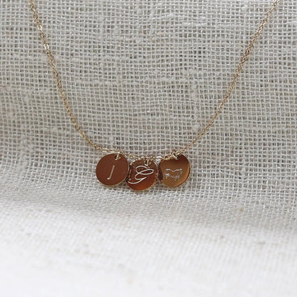 Collier "Inspiration" en acier inoxydable doré, composé de trois médaillons personnalisés d'initiales et dessin, présenté sur un fond beige texturé.