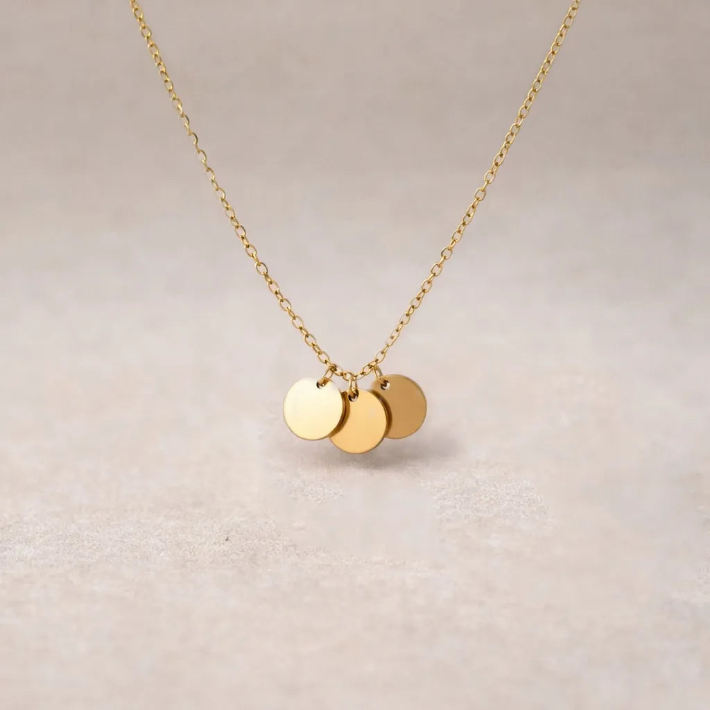 Collier "Inspiration" en acier inoxydable doré, composé de trois médaillons personnalisables, présenté sur un fond beige texturé.