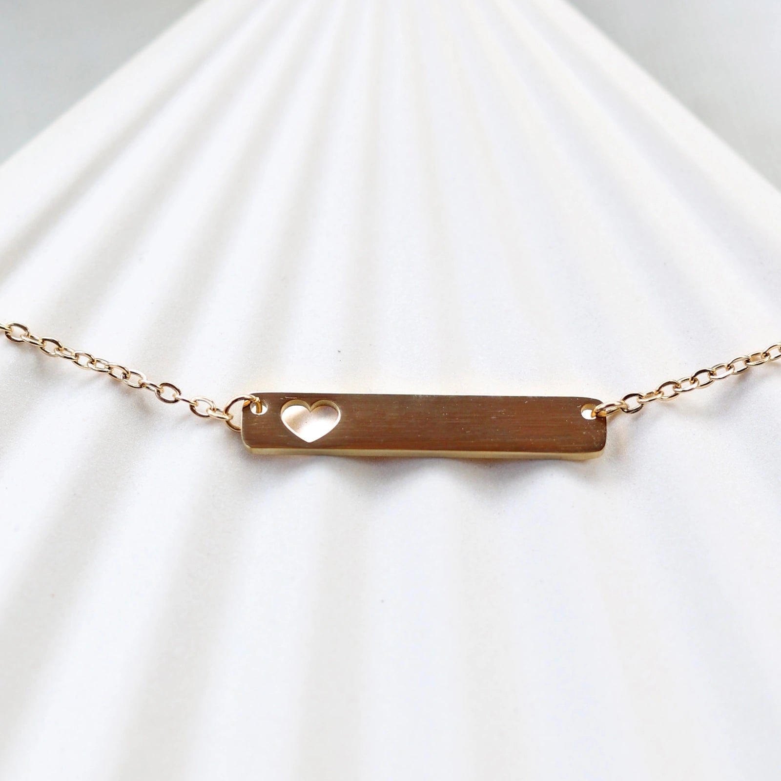 Collier plaque personnalisable en acier doré avec cœur découpé, posé à plat sur un support texturé beige, photo en gros plan. 