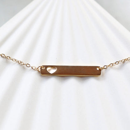 Collier plaque personnalisable en acier doré avec cœur découpé, posé à plat sur un support texturé beige, photo en gros plan. 