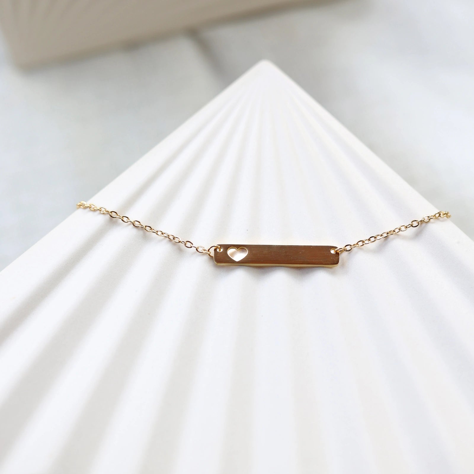 Collier plaque personnalisable en acier doré avec cœur découpé, posé à plat sur un support texturé beige, photo en lumière douce. 