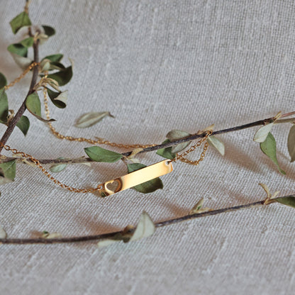 Zoom sur le collier "Touchante" avec plaque personnalisable en acier doré avec cœur découpé, posé sur une branche de feuilles
