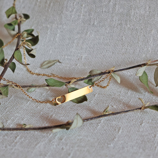 Zoom sur le collier "Touchante" avec plaque personnalisable en acier doré avec cœur découpé, posé sur une branche de feuilles