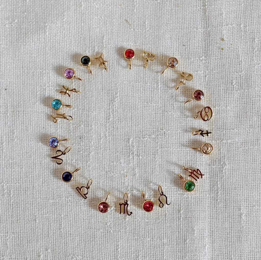 Vue d'ensemble des pendentifs "Mon identité" en acier doré, formant un cercle avec signes astrologiques et pierres colorées, posés sur un fond beige texturé.