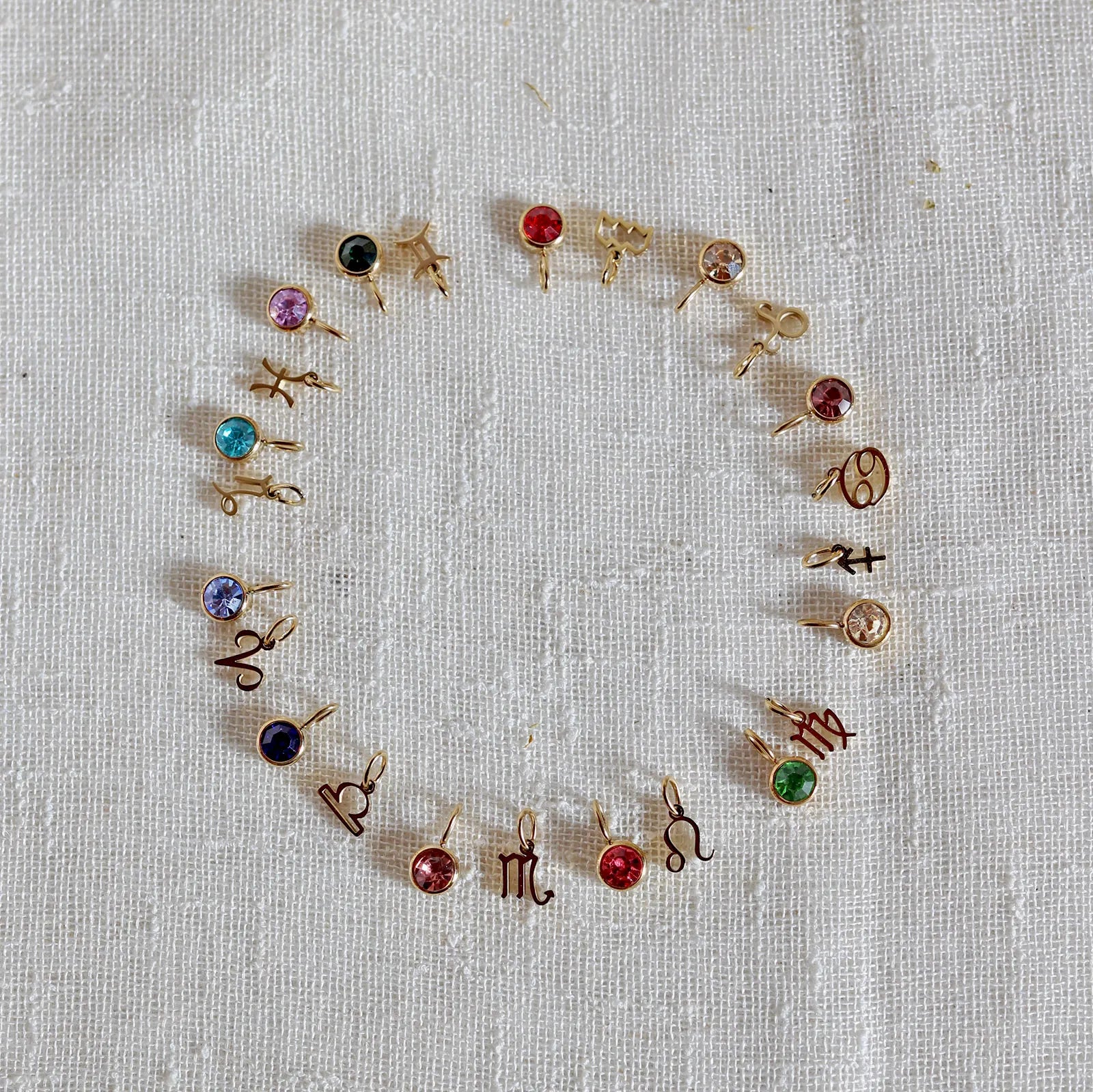 Vue d'ensemble des pendentifs "Mon identité" en acier doré, formant un cercle avec signes astrologiques et pierres colorées, posés sur un fond beige texturé.