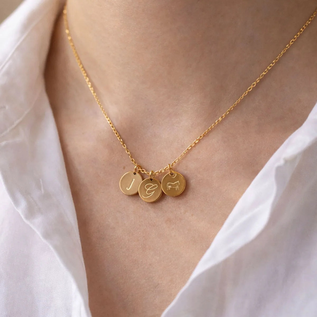 Collier "Inspiration" en acier inoxydable dorée, composé de trois médaillons personnalisables, porté autour du cou avec les initiales J G et symbole.