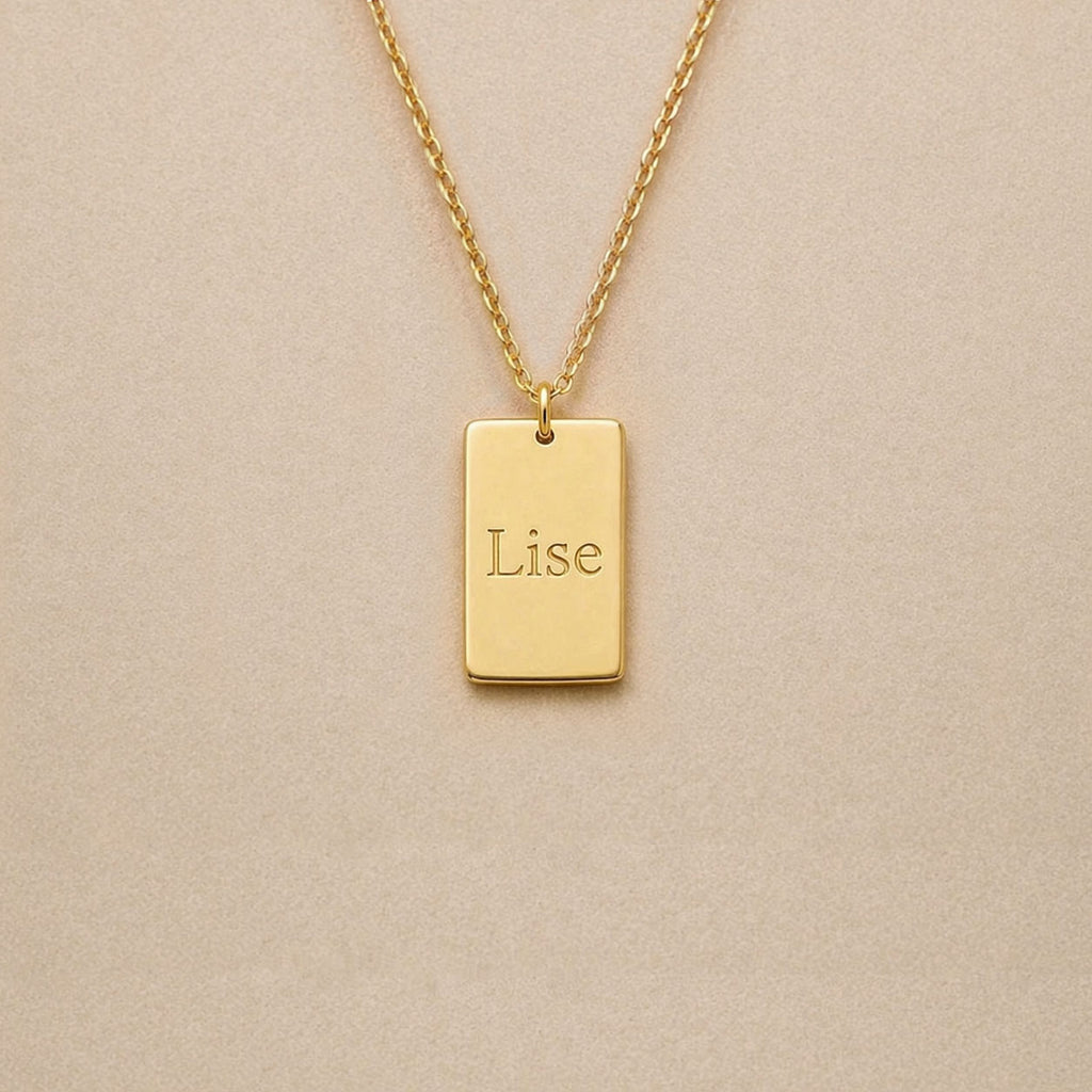 Collier Trace plaque rectangulaire personnalisable en acier inoxydable doré, gravé avec le prénom Lise, sur fond beige. 