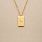 Collier Trace plaque rectangulaire personnalisable en acier inoxydable doré, gravé avec le prénom Lise, sur fond beige. 