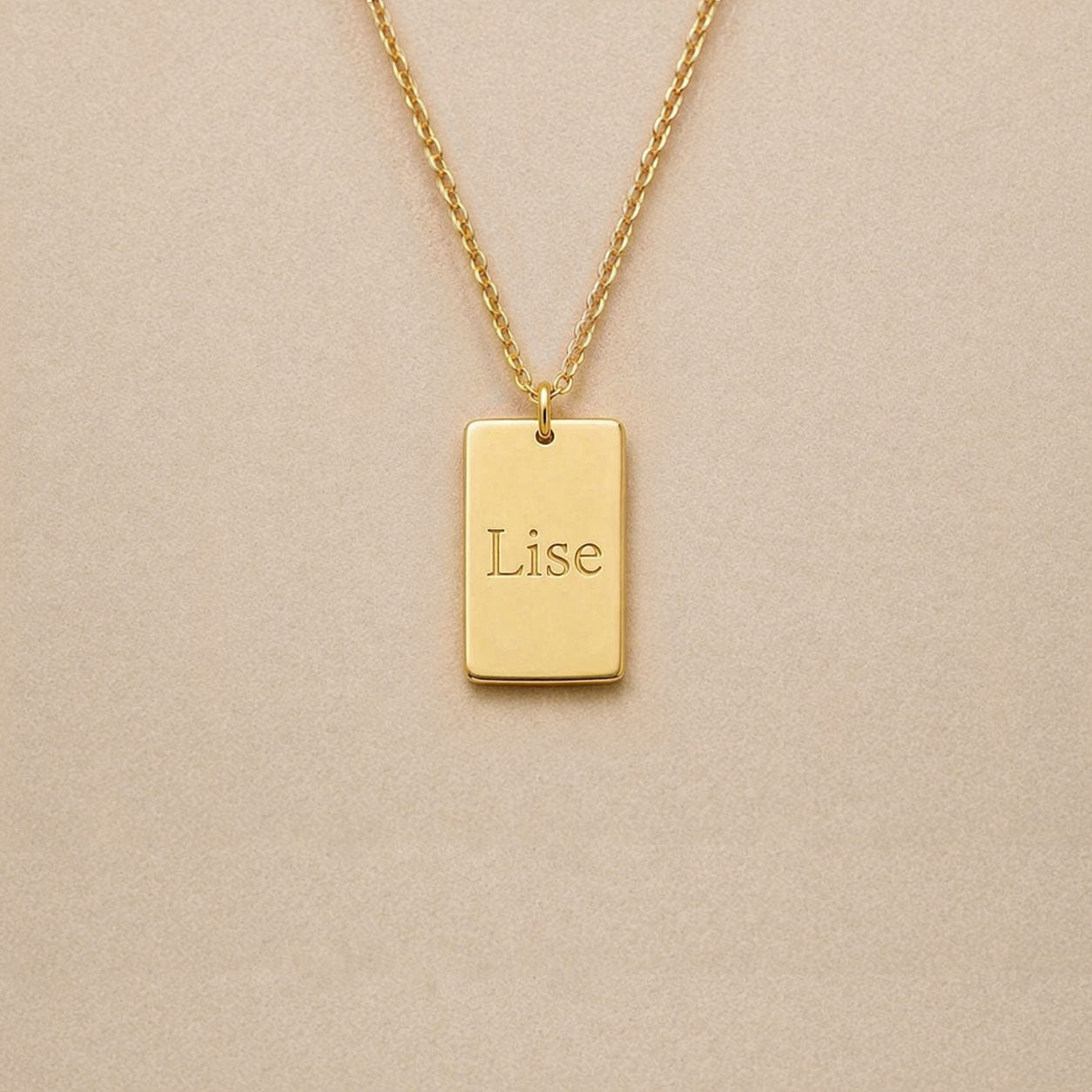 Collier Trace plaque rectangulaire personnalisable en acier inoxydable doré, gravé avec le prénom Lise, sur fond beige. 