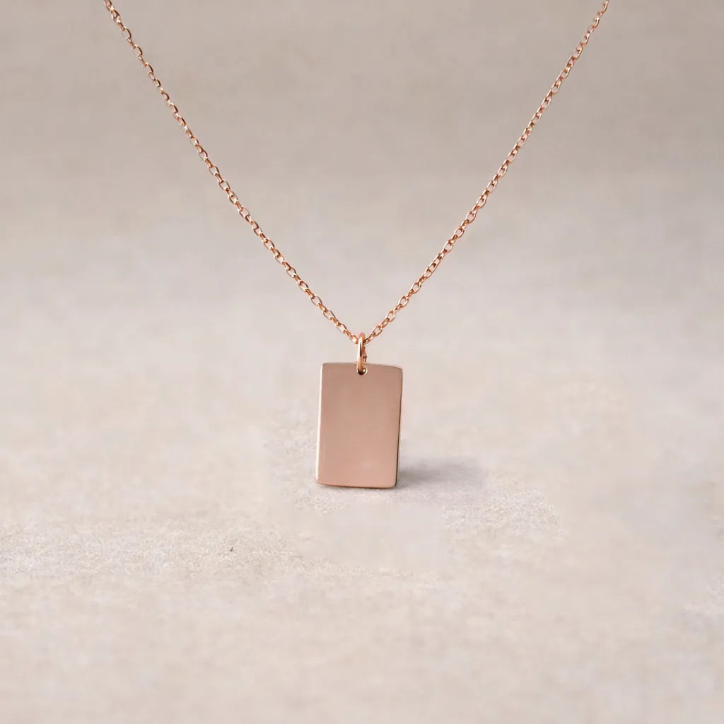 Zoom sur le collier "Je me souviens, en acier or rose, avec médaille rectangulaire à graver, posé sur un fond beige.