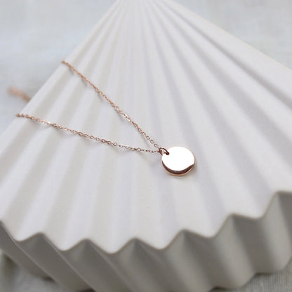 Collier personnalisable "Lumineuse", orné d'une médaille ronde brillante en acier or rose, posé sur un fond beige texturé.