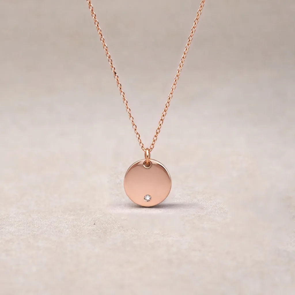 Zoom sur le collier personnalisable "Lumineuse" avec médaille ronde brillante en acier or rose, posé sur un fond beige texturé.