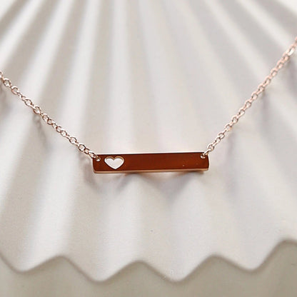 Collier plaque personnalisable en acier de couleur or rose avec cœur découpé, posé à plat sur un support texturé beige, photo en gros plan. 