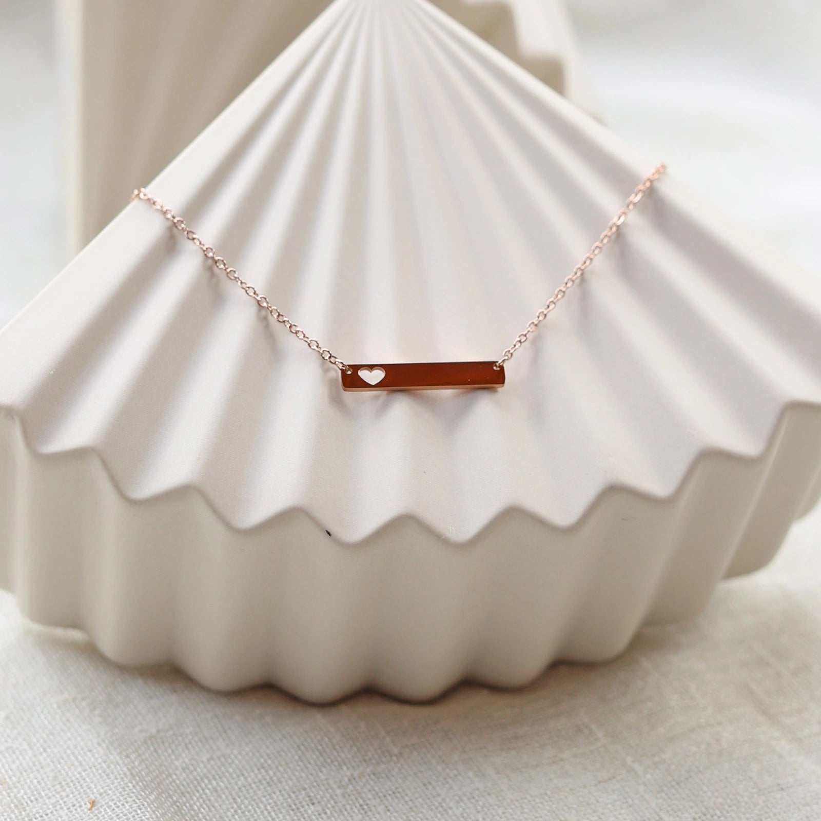 Collier plaque personnalisable en acier de couleur or rose avec cœur découpé, posé sur un support texturé beige, photo en lumière douce. 