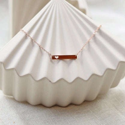 Collier plaque personnalisable en acier de couleur or rose avec cœur découpé, posé sur un support texturé beige, photo en lumière douce. 