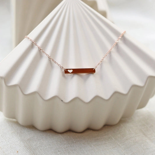 Collier plaque personnalisable en acier de couleur or rose avec cœur découpé, posé sur un support texturé beige, photo en lumière douce. 