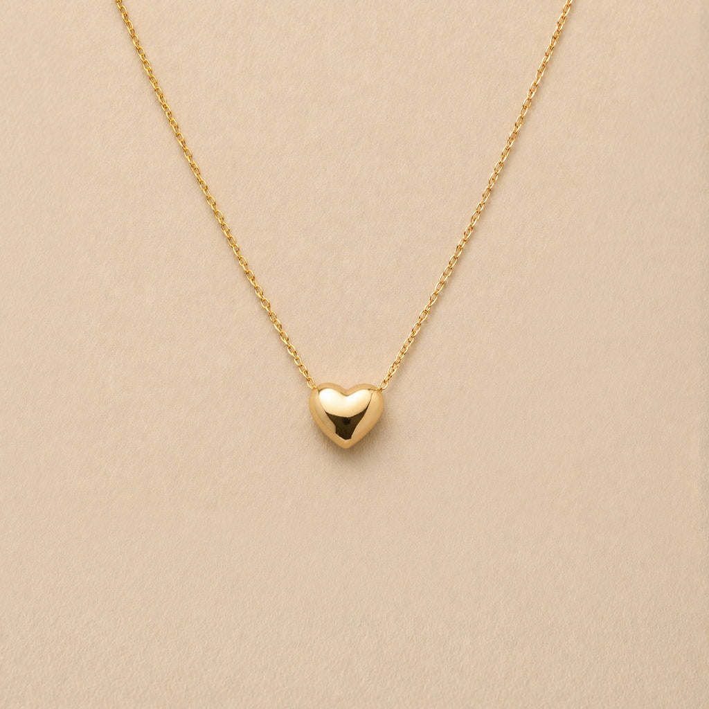 Collier Authentique avec pendentif cœur en acier inoxydable doré, photographié en gros plan sur un fond beige texturé.