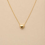 Collier Authentique avec pendentif cœur en acier inoxydable doré, photographié en gros plan sur un fond beige texturé.