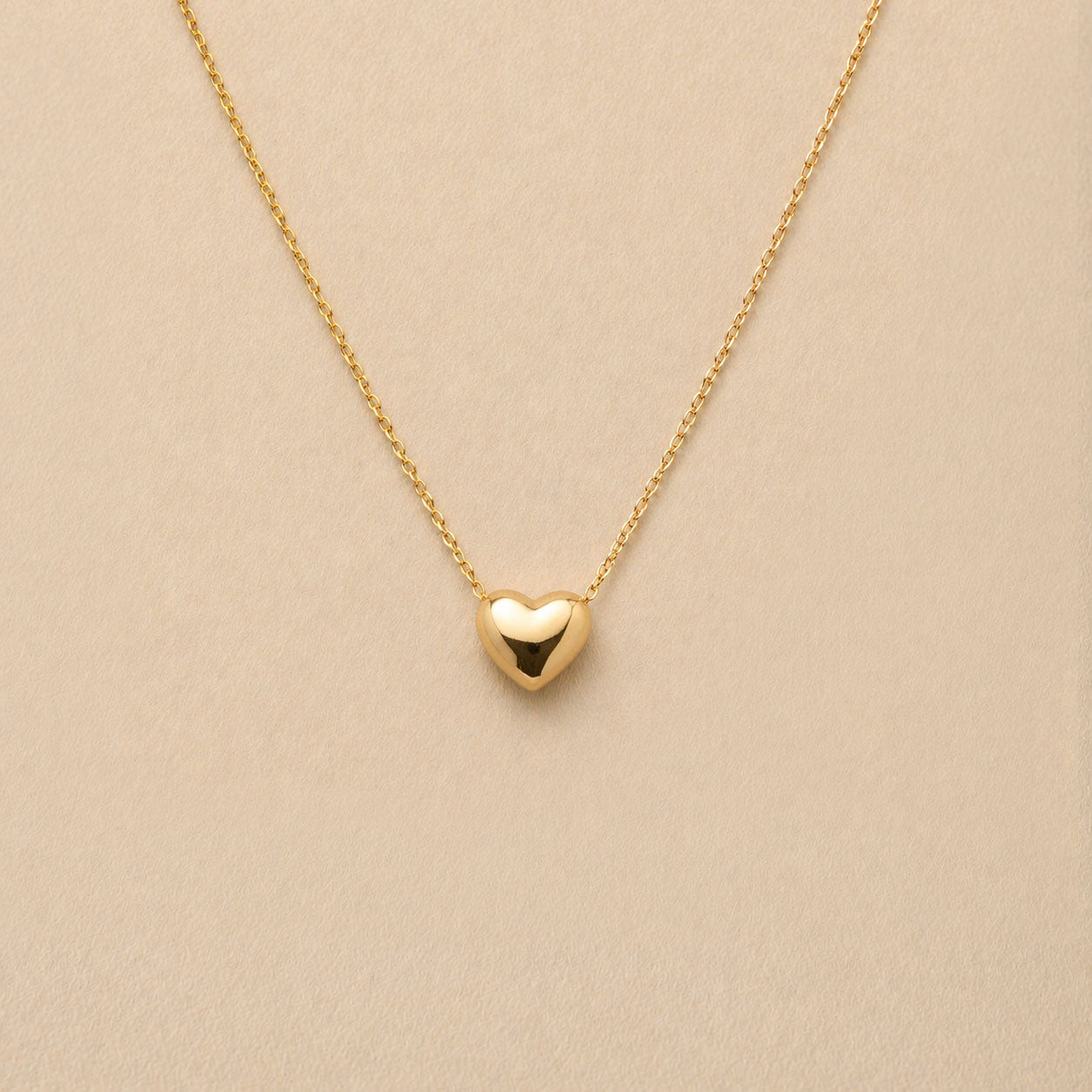 Collier Authentique avec pendentif cœur en acier inoxydable doré, photographié en gros plan sur un fond beige texturé.