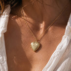 Zoom sur le collier Précieux, médaillon cœur en acier doré avec photo souvenir à personnaliser et gravure sur le devant du cœur,  posé sur un fond beige texturé.