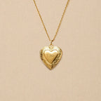 Zoom sur le collier Précieux, médaillon photo cœur en acier doré avec gravure prénom et  photo souvenir à personnaliser, posé sur un fond beige texturé.