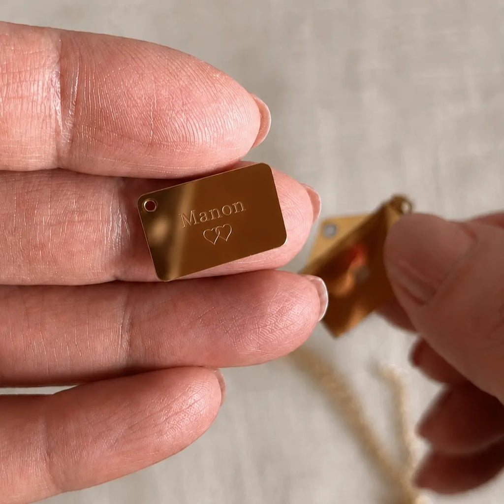  Collier "Nid secret", pendentif en forme d'enveloppe dorée ouverte, dévoilant un prénom Manon, posé dans la main.
