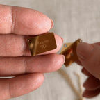  Collier "Nid secret", pendentif en forme d'enveloppe dorée ouverte, dévoilant un prénom Manon, posé dans la main.
