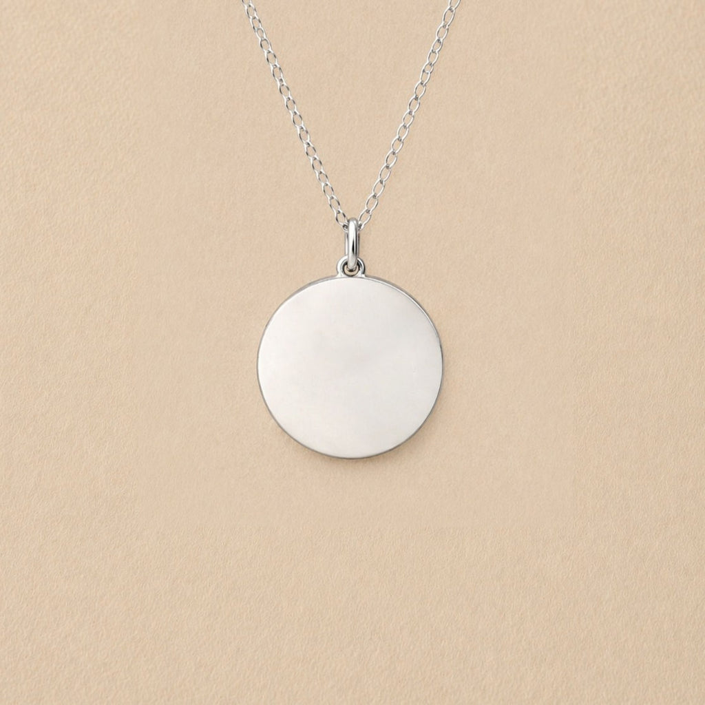 Collier "Reflet" avec médaille ronde personnalisé en acier inoxydable argenté, posé sur un fond beige texturé