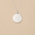 Collier "Reflet" avec médaille ronde personnalisé en acier inoxydable argenté, posé sur un fond beige texturé